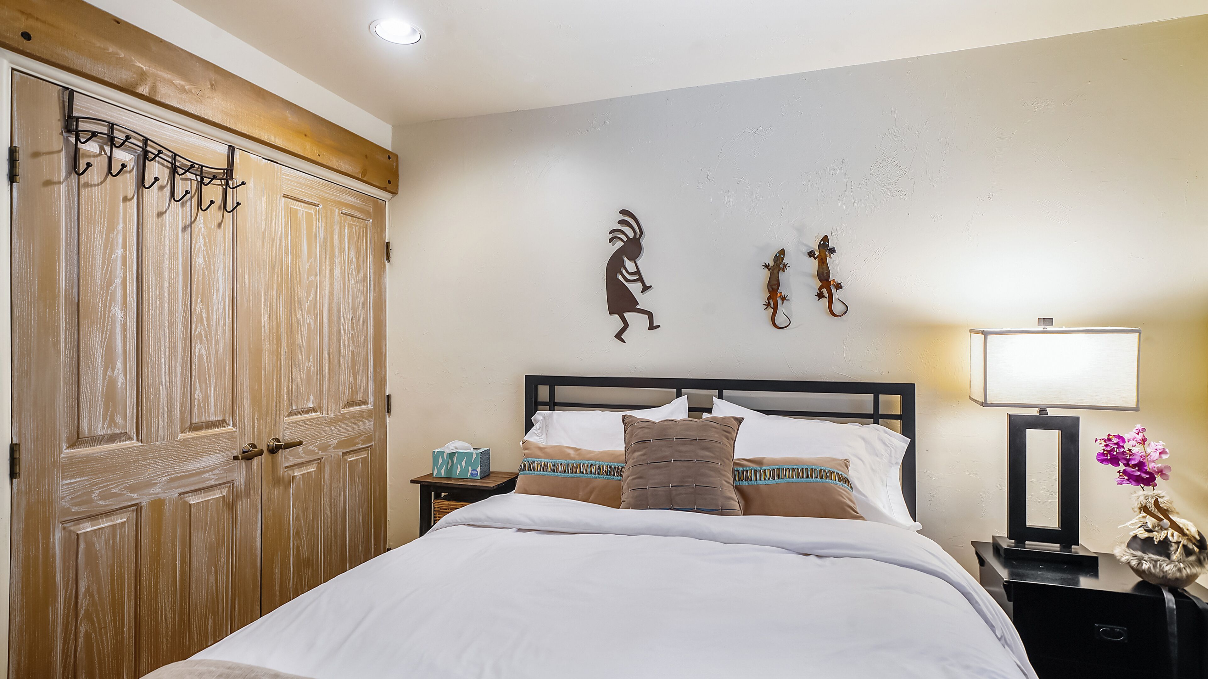 A bedroom with a wooden wardrobe, a bed with multiple pillows, a bedside table with a lamp, and wall art.