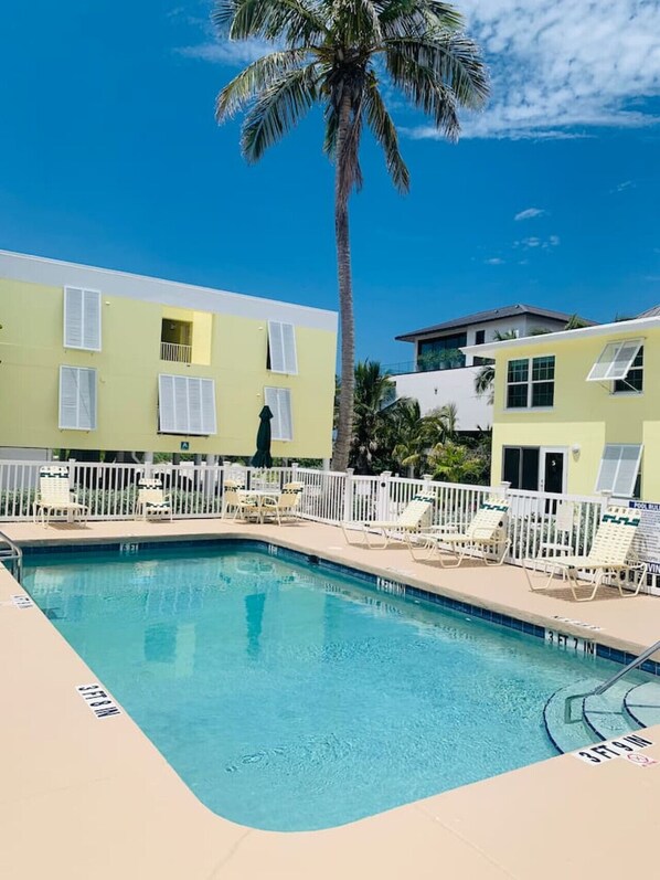 A pool area with lounge chairs and a palm tree, surrounded by buildings with balconies. - Cabana Beach Club - Beachfront Stunning Ocean View *Snow Bird Promo* Winter 2027 (Longboat Key)