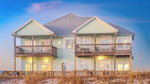 A two-story beachfront house with a balcony on each floor, a wooden deck, and a sandy beach in the foreground.