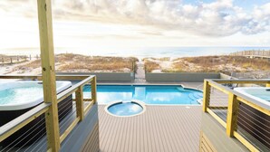 A wooden deck with a pool and hot tubs overlooking a beach.