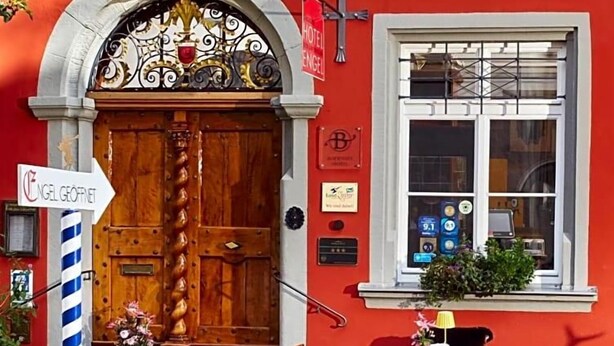 A red building with a wooden door, a sign for Hotel Engel, outdoor seating with tables and chairs, and a rope barrier. - Hotel Engel Lindauer Bier Und Weinstube (Lindau)