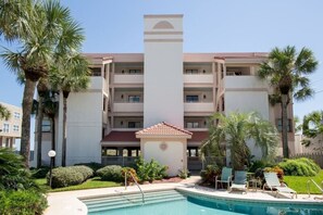 A multi-story hotel with a swimming pool, palm trees, and a covered area with chairs.