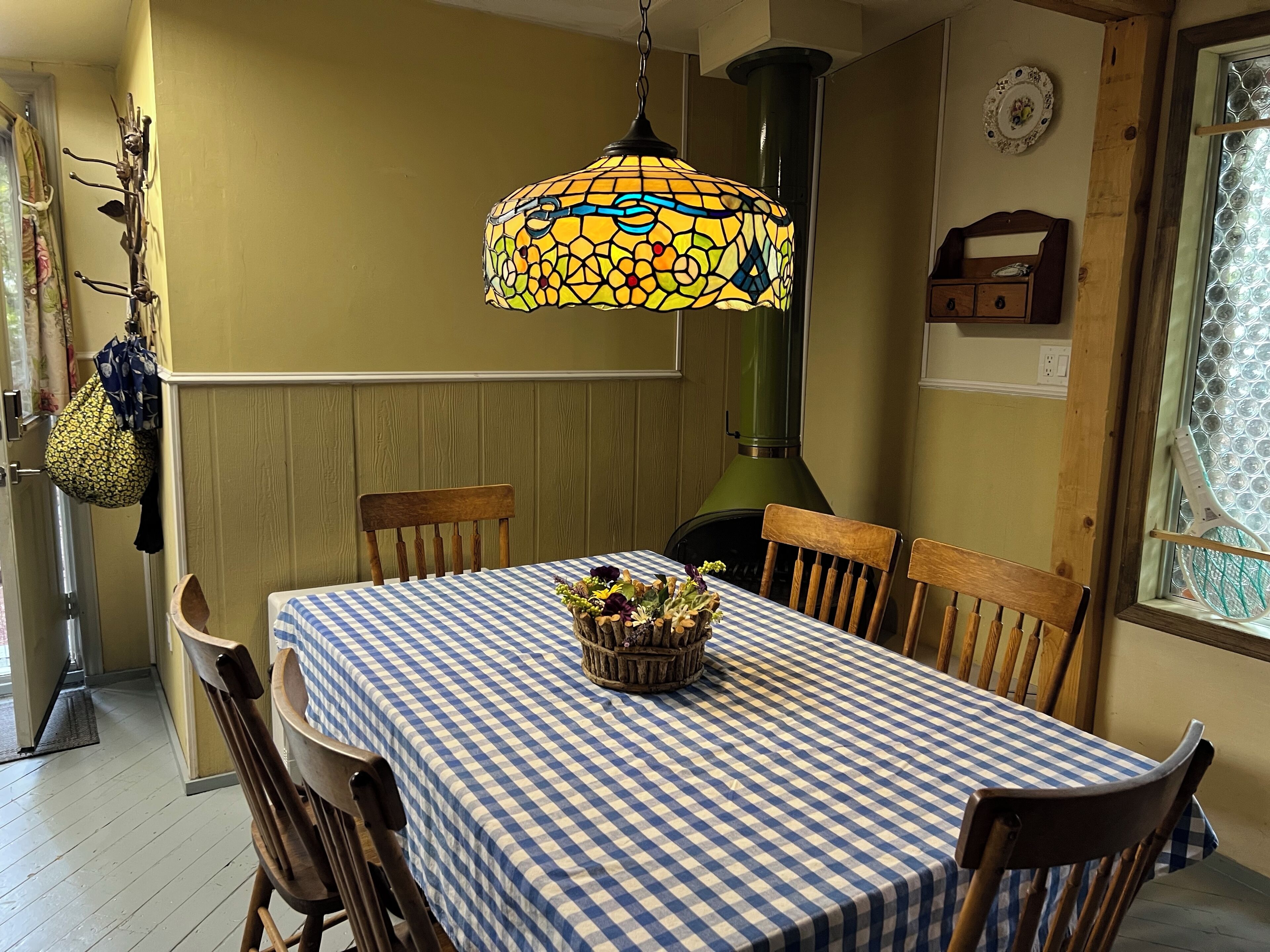 Un comedor con mantel a cuadros, sillas de madera, una lámpara colgante y un ventanal con celosía decorativa.