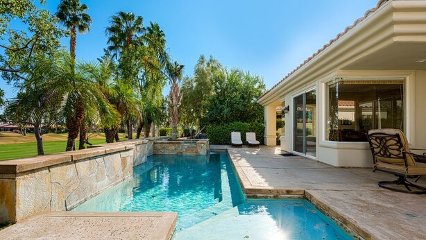A swimming pool with a stone border, a patio area with a chair, and palm trees in the background.