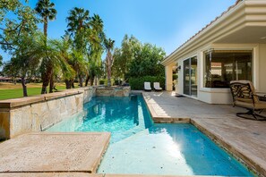 Una piscina con borde de piedra, un patio con una silla y palmeras al fondo.