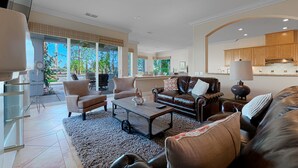 A spacious living room with a leather sofa, a coffee table, and a TV. There is a kitchen area visible in the background.