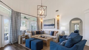 A living room with a sofa, ottomans, a coffee table, a chandelier, a painting, and a window with curtains. - Blue Haven, Tivoli, Sandestin, Golf Cart (Miramar Beach)