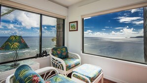 Een kamer met een groot raam met uitzicht op zee en lucht. Er is een stoel met een bloemenkussen, een kleine tafel met een lamp, en een ingelijst kunstwerk aan de muur.
