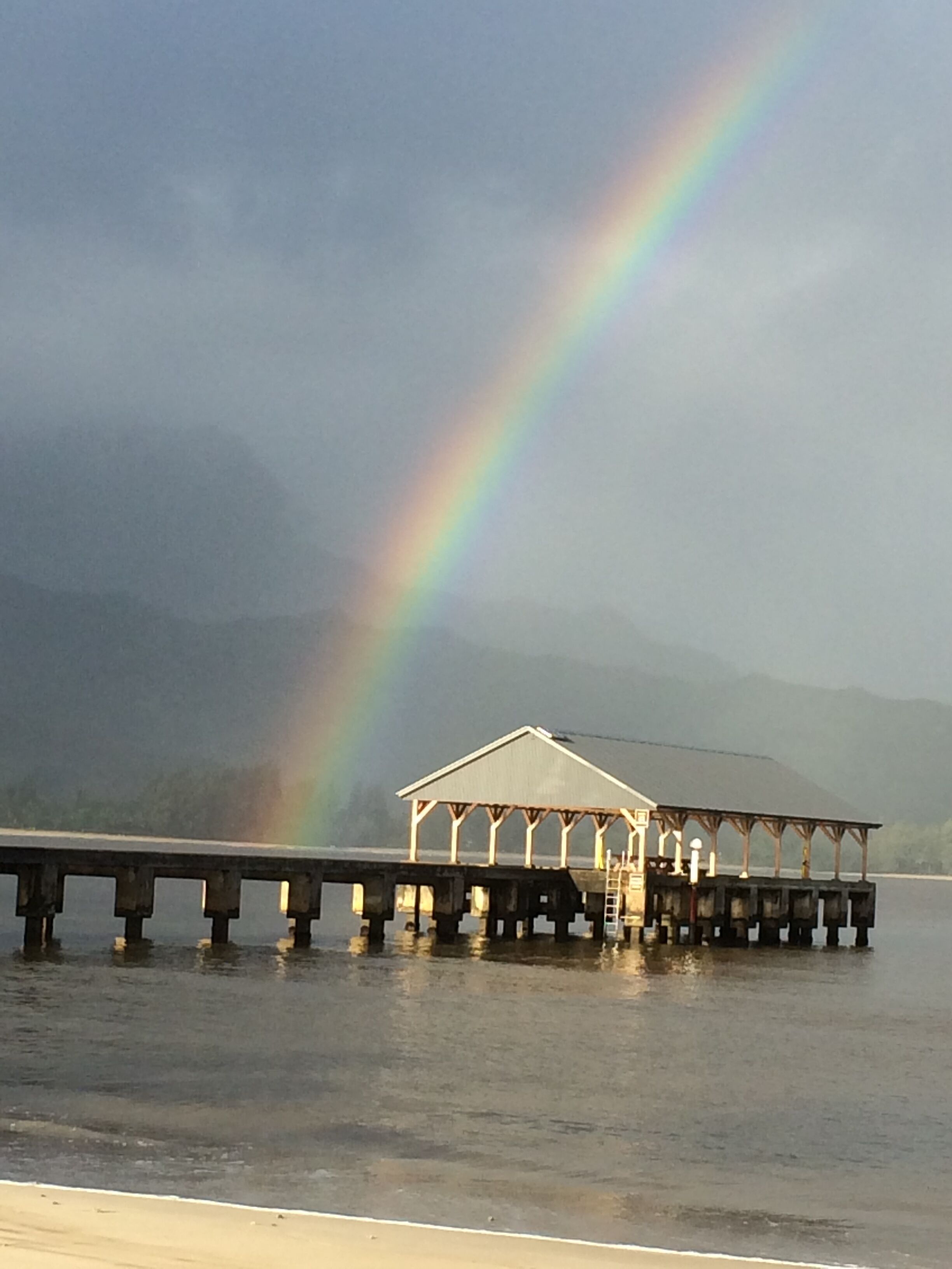 A pier with a covered structure, extending over calm water with a rainbow in the background.
