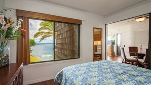 Een slaapkamer met een groot raam met uitzicht op de oceaan, een bed met een bloemenlaken, een houten bureau met een bloevas en een eetgedeelte met rieten stoelen.