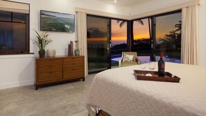 A bedroom with a bed, a wooden dresser, a TV, and a view of a sunset over a pool. - Ocean Views/Private Swimming Pool. Mauna Kea High Bluffs Home (Mauna Kea Beach Resort)