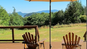A deck with two wooden Adirondack chairs and a large umbrella, overlooking a grassy area with trees and distant mountains. - ⚓️ Nautical Sea Cabin with Water View ⚓️ (Lopez Island)