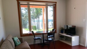 Een woonkamer met een bank, een eettafel met stoelen, een televisie op een standaard en een groot raam met uitzicht op bomen en een wateroppervlak.