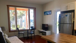 Een knus woongedeelte met een kitchenette, een eethoek met tafel en stoelen, een bank met een bloemenkussen, en een televisie op een standaard.