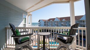 Un balcone con due poltrone Adirondack e un tavolino, con vista su una piscina e un edificio fronte spiaggia.