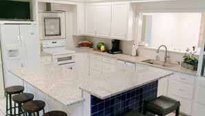 Eine moderne Küche mit weißen Schränken, einer Granit-Arbeitsplatte, einer eingebauten Kochinsel mit Hockern, einer Kaffeemaschine und einem Spülbecken.