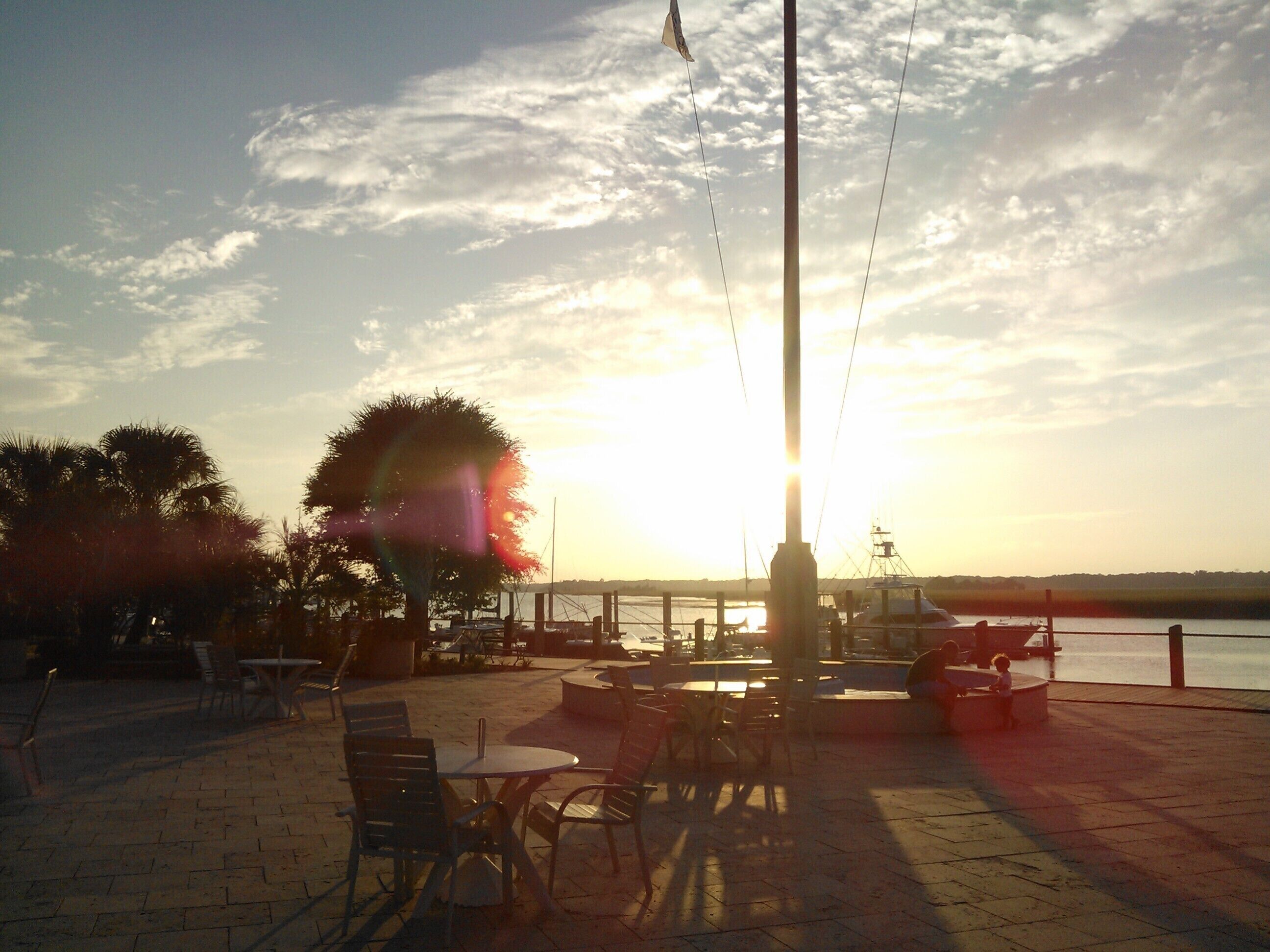 An outdoor seating area with tables and chairs, a flagpole, and a view of the water and sunset.