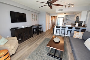 A modern living room with a sofa, coffee table, and TV. It includes a kitchen with bar stools and a dining area.
