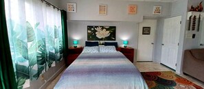 Um quarto com uma cama, duas mesas de cabeceira com luminárias, um quadro floral grande na parede e uma porta com espelho.