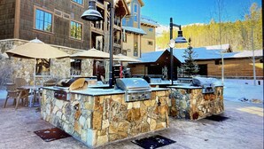 Zona de bar al aire libre con mostradores de piedra y estaciones de parrillada, rodeada de edificios de madera y un paisaje nevado.
