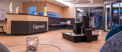 A modern reception area with a wooden reception desk labeled 'Lipprandt', a seating area with a sofa and chairs, and a decorative water feature.
