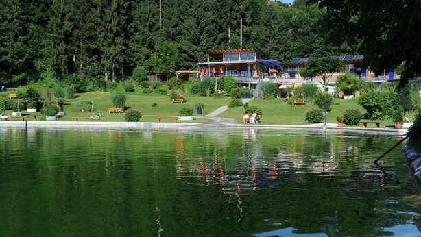 A lakeside hotel with a swimming area, outdoor seating, and a view of the forested hills.