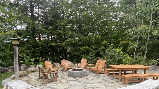 Un foyer en pierre, entouré de fauteuils Adirondack en bois et d’une table, est installé sur une terrasse en pierre, agrémentée d’un muret en pierre et d’un lampadaire.