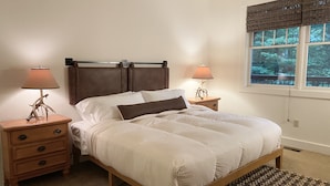Une chambre à coucher avec un lit, deux tables de chevet, une fenêtre avec des stores et une vue sur la verdure à l’extérieur.