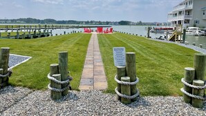 A waterfront property with a grassy lawn, a pathway leading to red chairs, and a sign indicating 'Bethany Marina Phase II Welcome to the Point'.