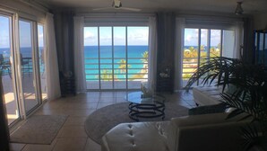 A spacious room with a large sliding glass door leading to a balcony with a view of the ocean. There is a round glass table with a chair, a sofa, and a potted plant. - Oceanfront Spectacular Penthouse W/Spacious Rooftop Terrace, Cupecoy Beach (Cupecoy)