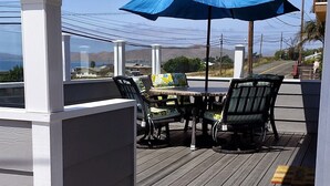 A wooden deck with outdoor furniture, including a table and chairs, under a blue umbrella. - FULL OCEAN VIEW!!! 3 minute walk to beach, WiFi, Dogs welcomed upon approval (Cayucos)