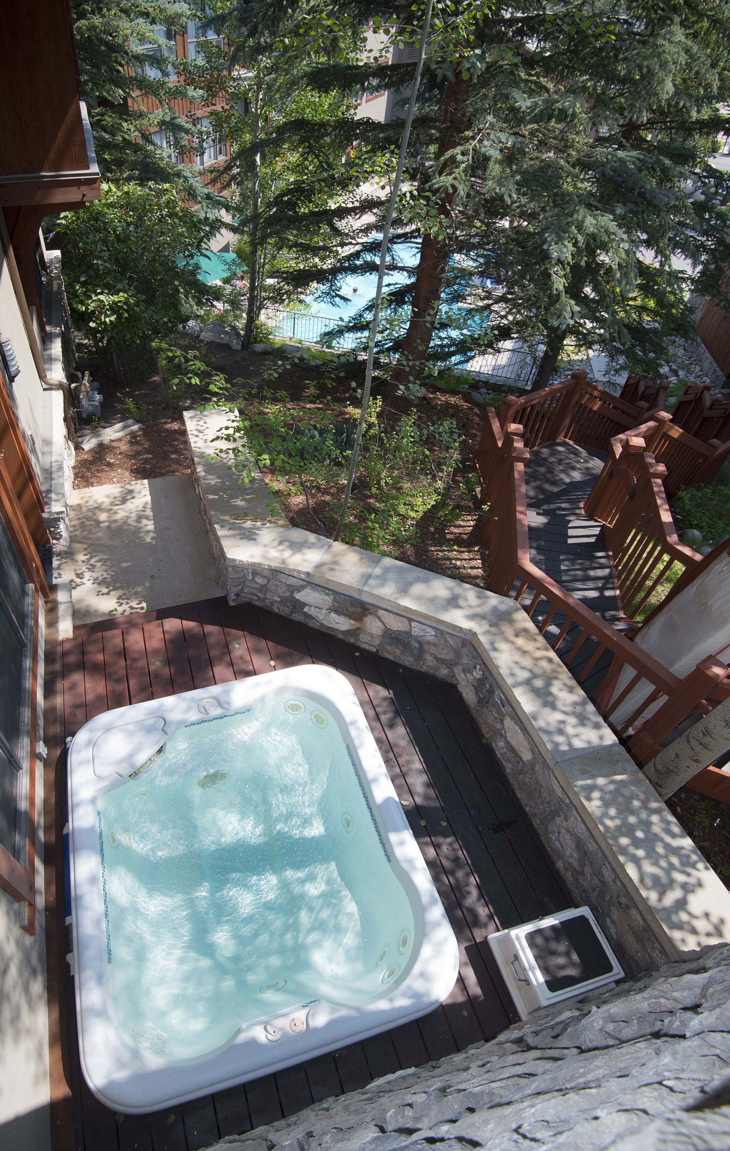 Tinaco en una terraza de madera con borde de piedra, rodeado de árboles y vista a una piscina.