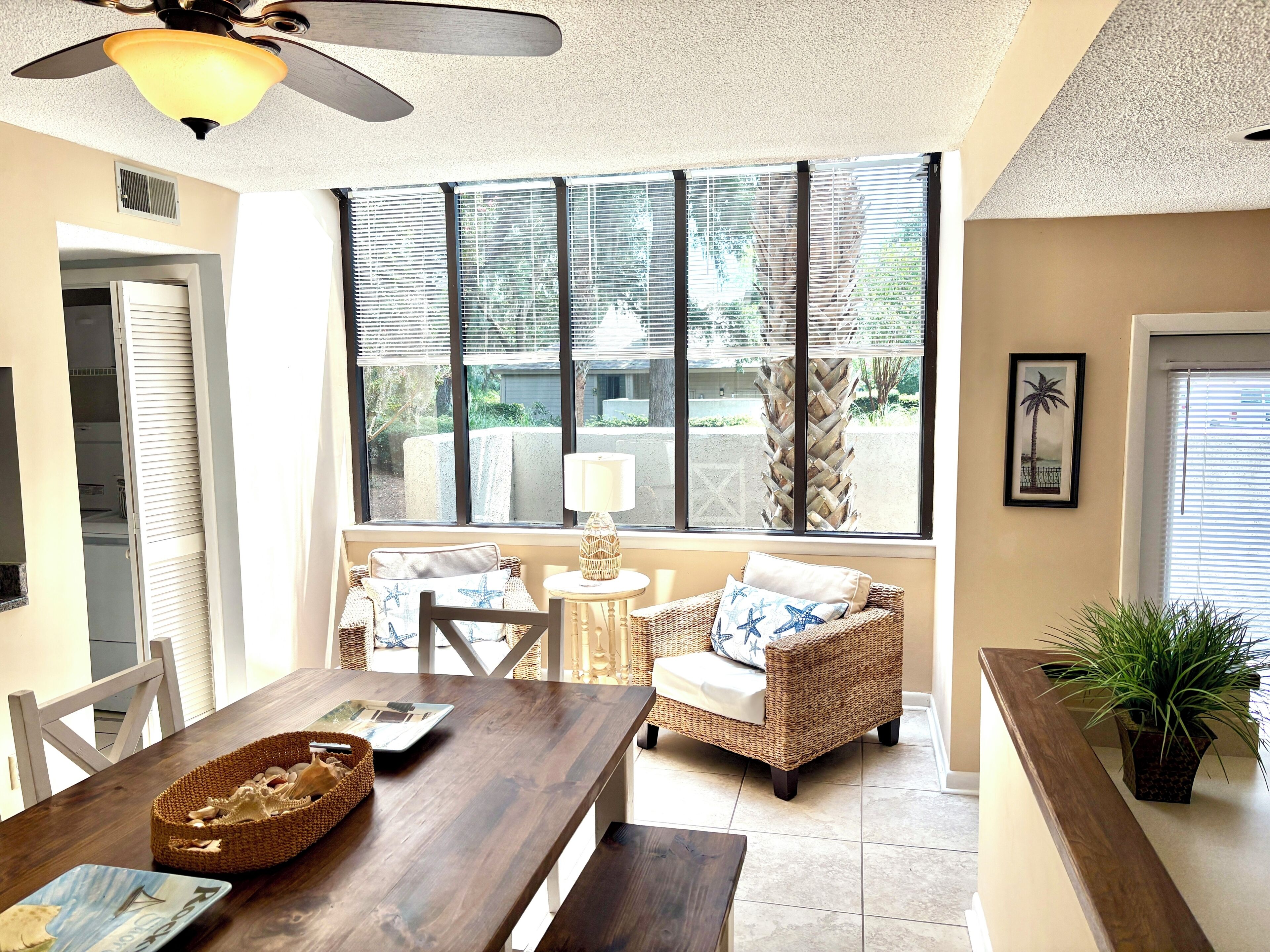 A sunlit room with a dining table, wicker chairs, a ceiling fan, and a view of the outdoors through large windows.