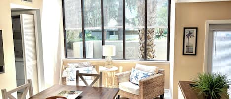 A sunlit room with a dining table, wicker chairs, a ceiling fan, and a view of the outdoors through large windows.