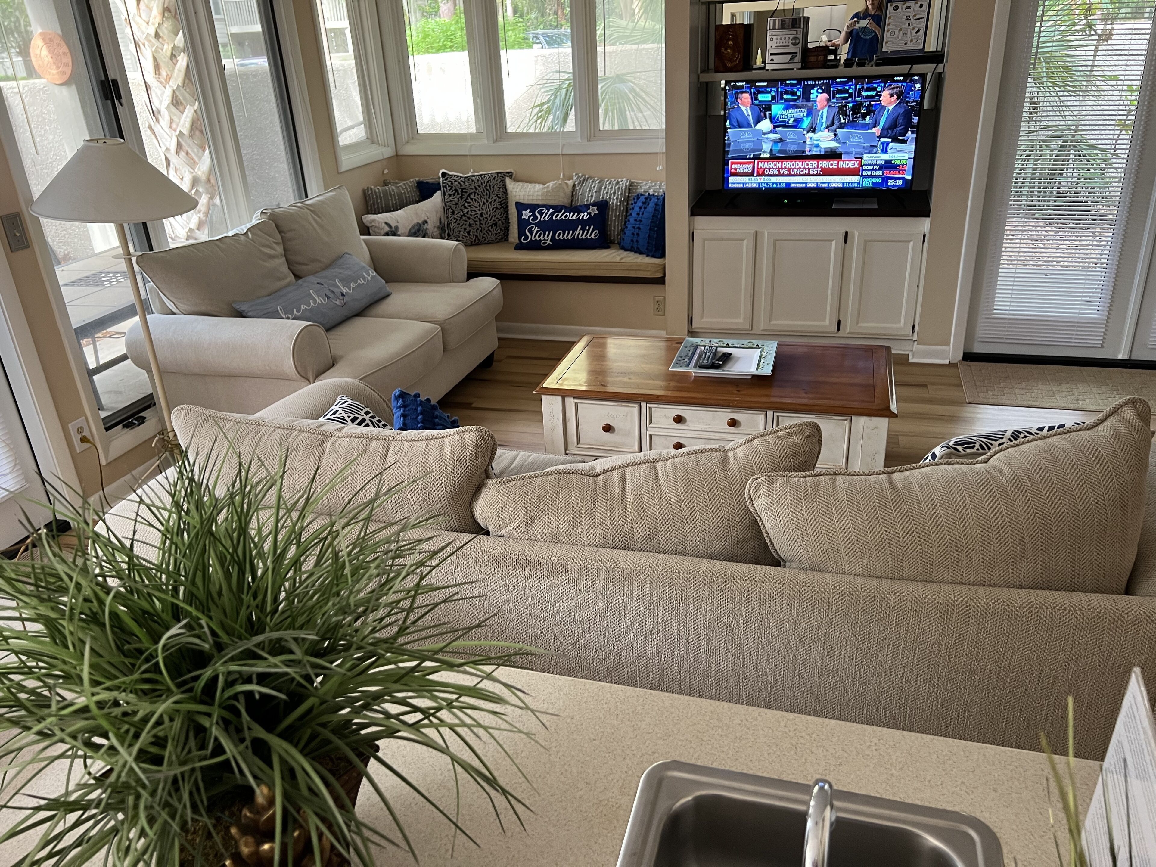 A living room with a beige sofa, a coffee table, and a television displaying a news program.