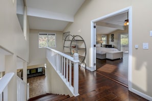 A spacious room with a high ceiling, a wooden staircase, a white railing, a metal shelf with a bowl, and a view of another room with a bed and a window.