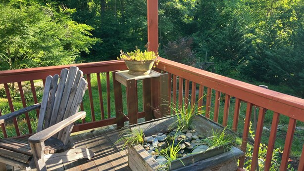 Una terraza de madera con barandal rojo, una silla de madera y una jardinera con plantas y rocas.