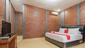 A bedroom with a brick-patterned wall, a bed with white bedding and red accents, a wooden desk with a television, and a tiled floor. - RedDoorz Plus @ Ampera Raya (Jakarta)