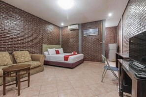 Habitación de hotel con pared de ladrillo, cama con ropa blanca y detalles rojos, sofá, mesita, escritorio con computadora y una silla.