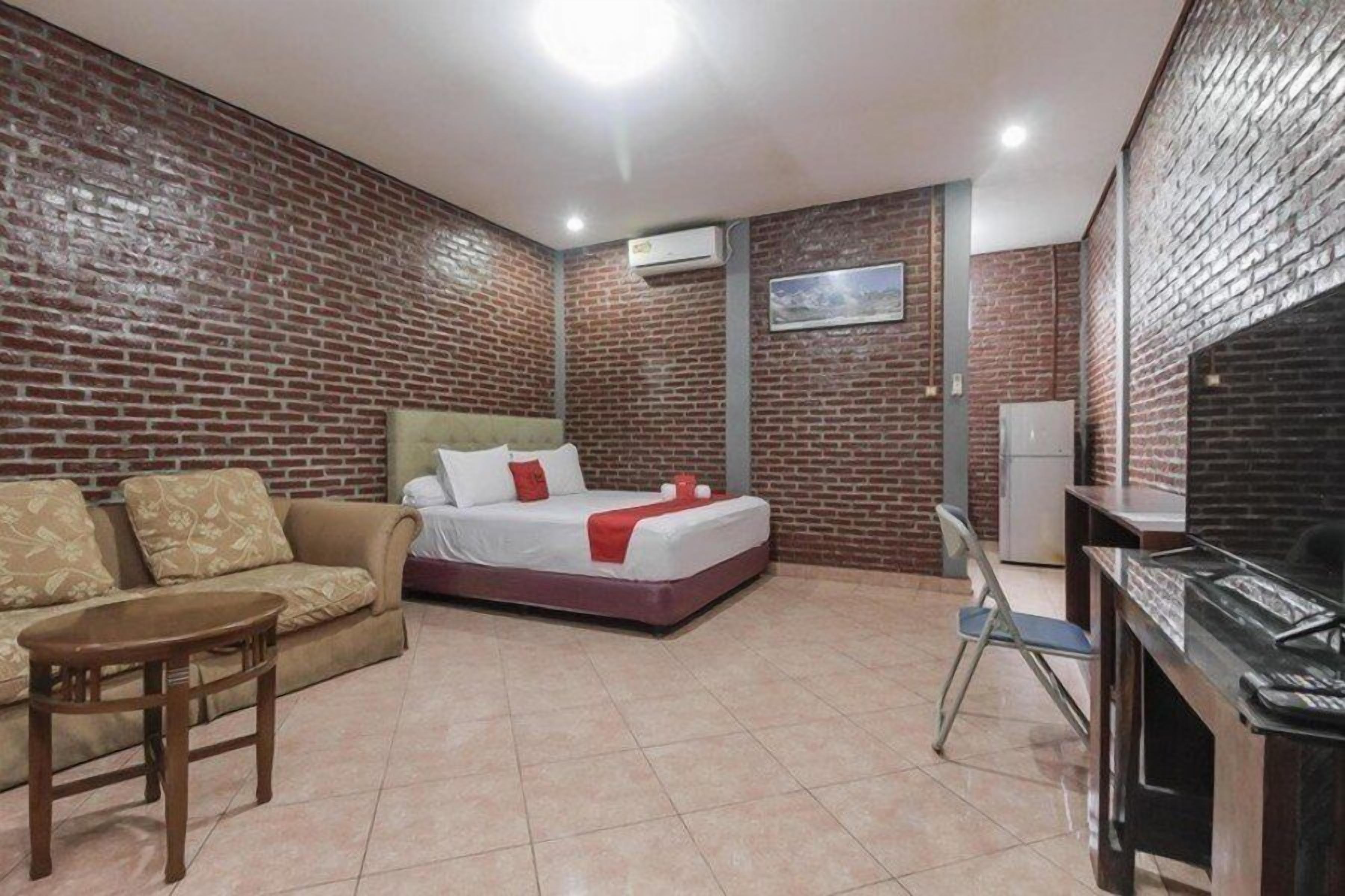 Ein Hotelzimmer mit Backsteinwand, einem Bett mit weißer Bettwäsche und roten Akzenten, einer Couch, einem kleinen Tisch und einem Schreibtisch mit einem Computer.