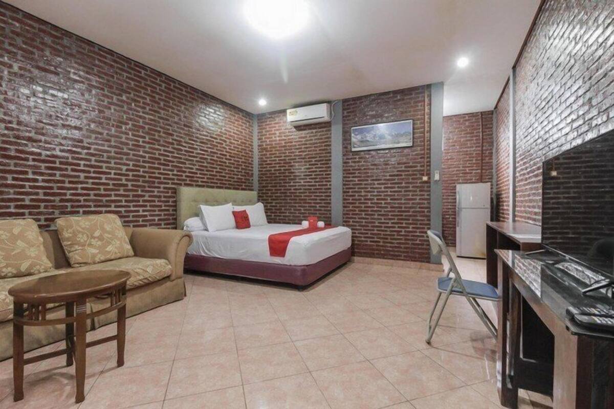 A hotel room with a brick wall, a bed with white bedding and red accents, a sofa, a small table, and a desk with a computer.
