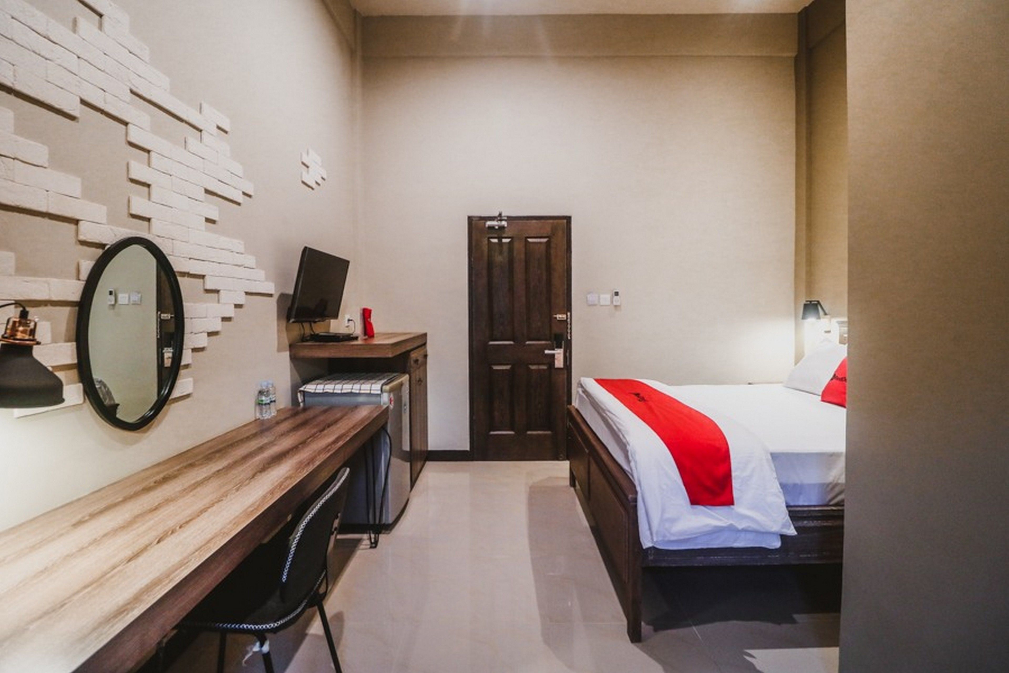 A compact hotel room with a wooden desk, a bed with red bedding, a mirror, a television, and a door.