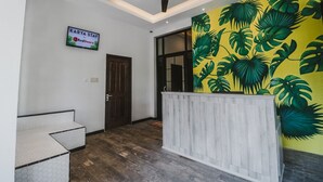 Un espace d’accueil doté d’un plancher en bois, d’un ventilateur de plafond, d’une enseigne indiquant « KARYA STAY » et d’un mur orné d’une fresque représentant des feuilles tropicales.