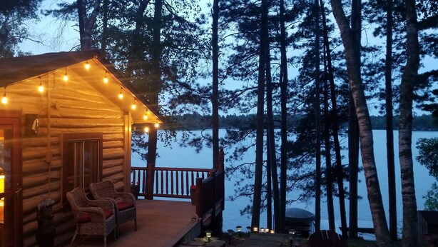 A log cabin with a porch, wicker chairs, and a view of a lake surrounded by trees. - Rustic Log Cabin/Modern Addition - Pontoon, Bunk House, Hot Tub - Woman Chain (Longville)