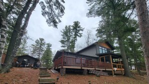A cabin with a wooden deck, stone steps, and a view of surrounding trees. - Rustic Log Cabin/Modern Addition - Pontoon, Bunk House, Hot Tub - Woman Chain (Longville)