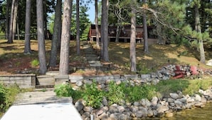 A cabin with a wooden deck, surrounded by trees and a small stream. - Rustic Log Cabin/Modern Addition - Pontoon, Bunk House, Hot Tub - Woman Chain (Longville)