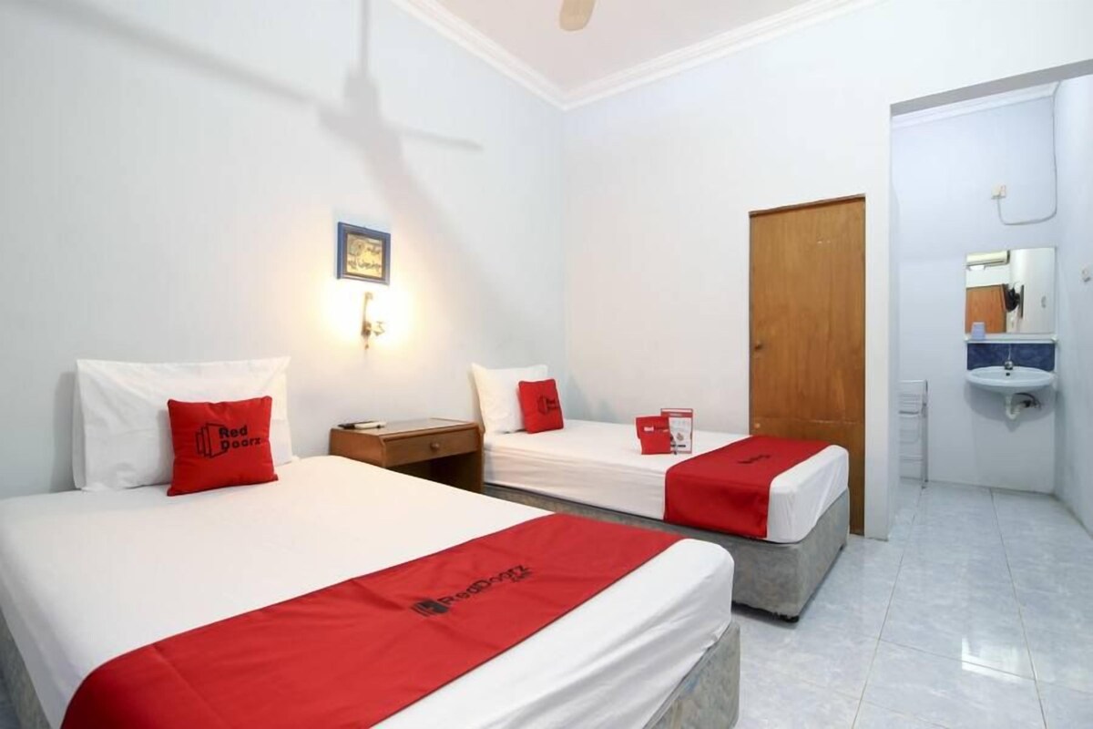 Habitación de hotel con dos camas, cada una con colcha y almohadas rojas a juego. Hay una mesita de noche de madera entre las camas, una pequeña mesa con una lámpara, y un aplique en la pared. Una puerta conduce a un baño con lavabo y espejo.