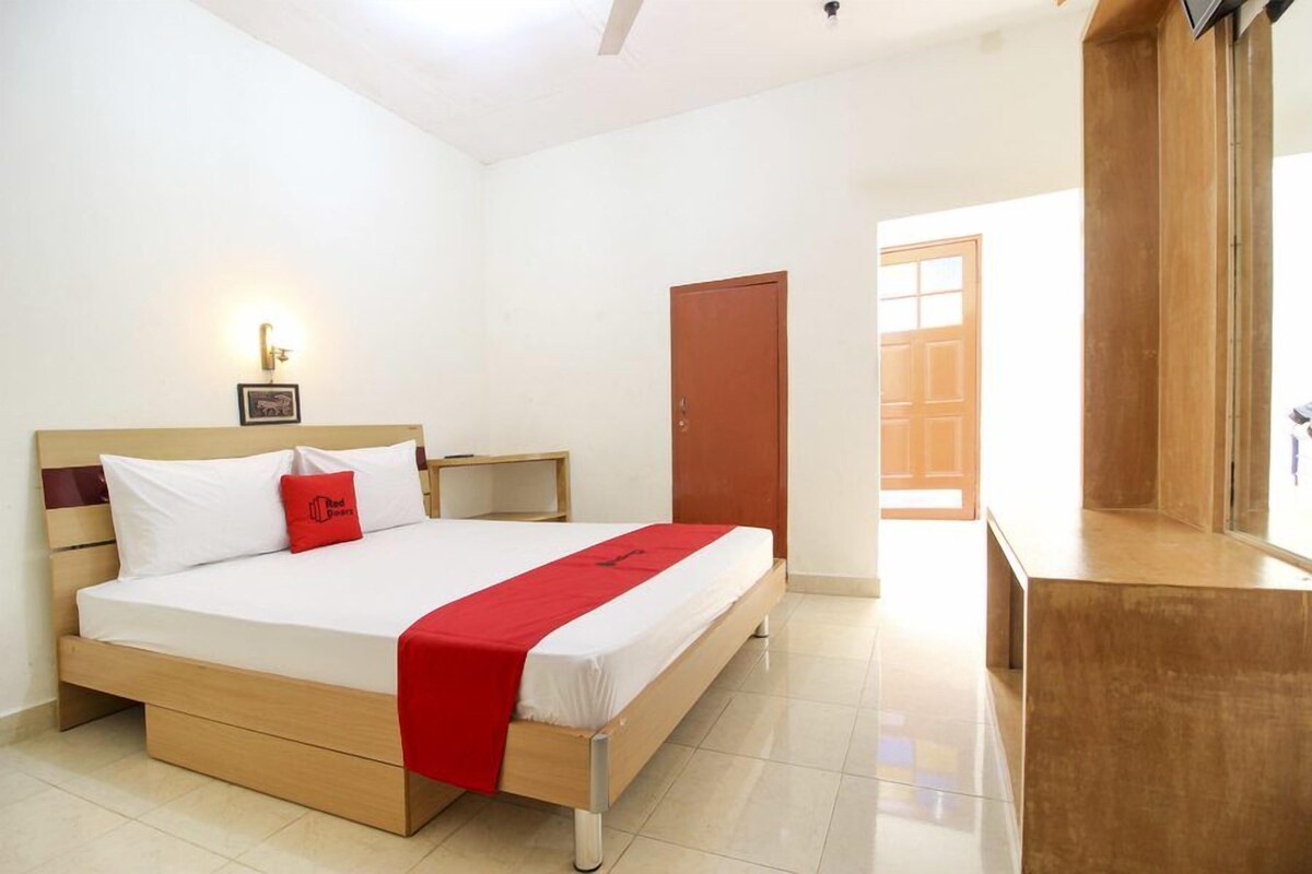 Habitación de hotel con una cama, una mesita de noche de madera, un almohadón rojo, una colcha blanca y una puerta de madera.