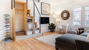 Ein modernes Wohnzimmer mit Treppe, einem Flachbildfernseher, einem Schreibtisch mit Stuhl, einem grauen Sofa und einem Bücherregal voller Bücher.