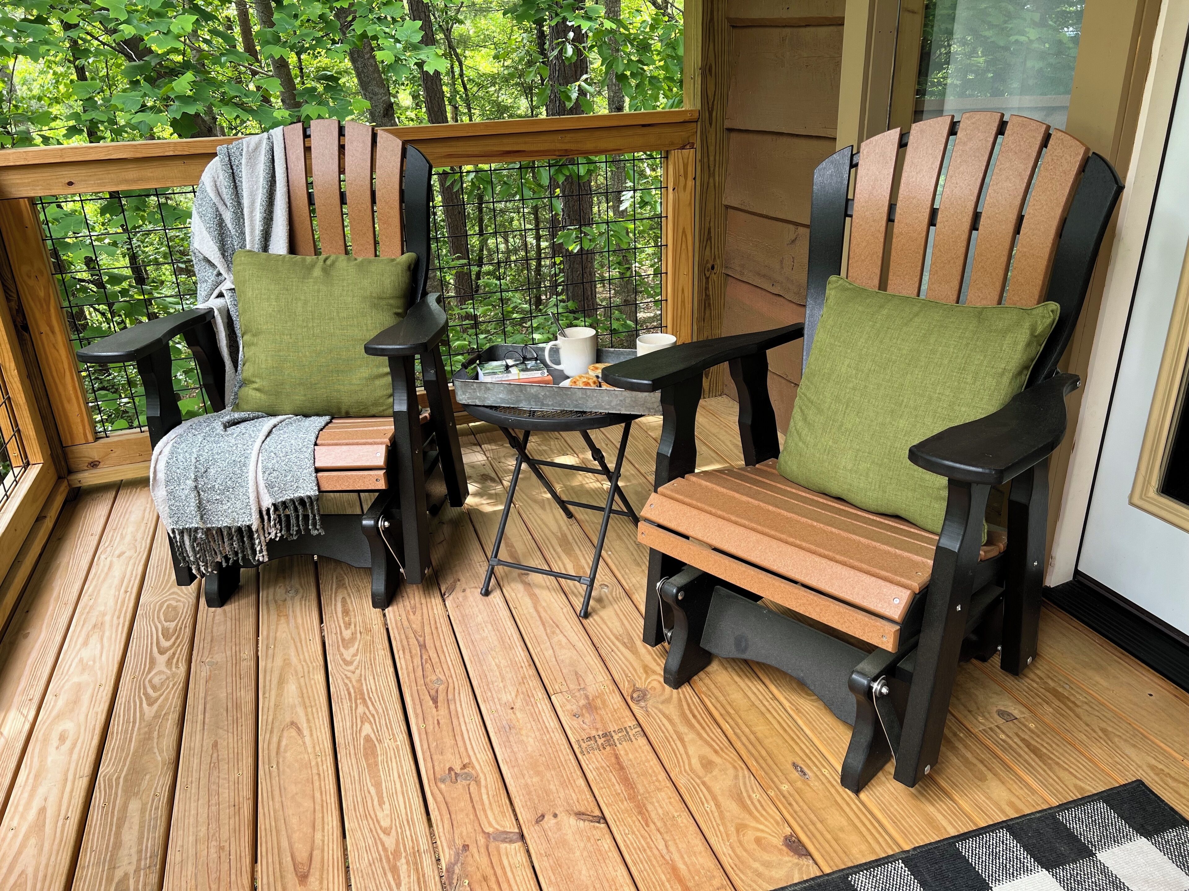A wooden deck with two Adirondack chairs and a small table, surrounded by a wooden railing and a glass door.